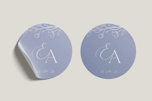 Two versions of a circular wedding logo sticker featuring the initials "E & A" with floral accents and the date "14 | 09 | 24" on a lavender-blue background, one of which appears to be peeling at the edge