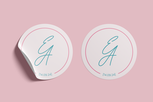 Two versions of a circular wedding sticker featuring the initials "E & A" in the teal script, a pink border, and the date "14.09.24" beneath, set against a blush pink background, with one sticker appearing to peel at the edge