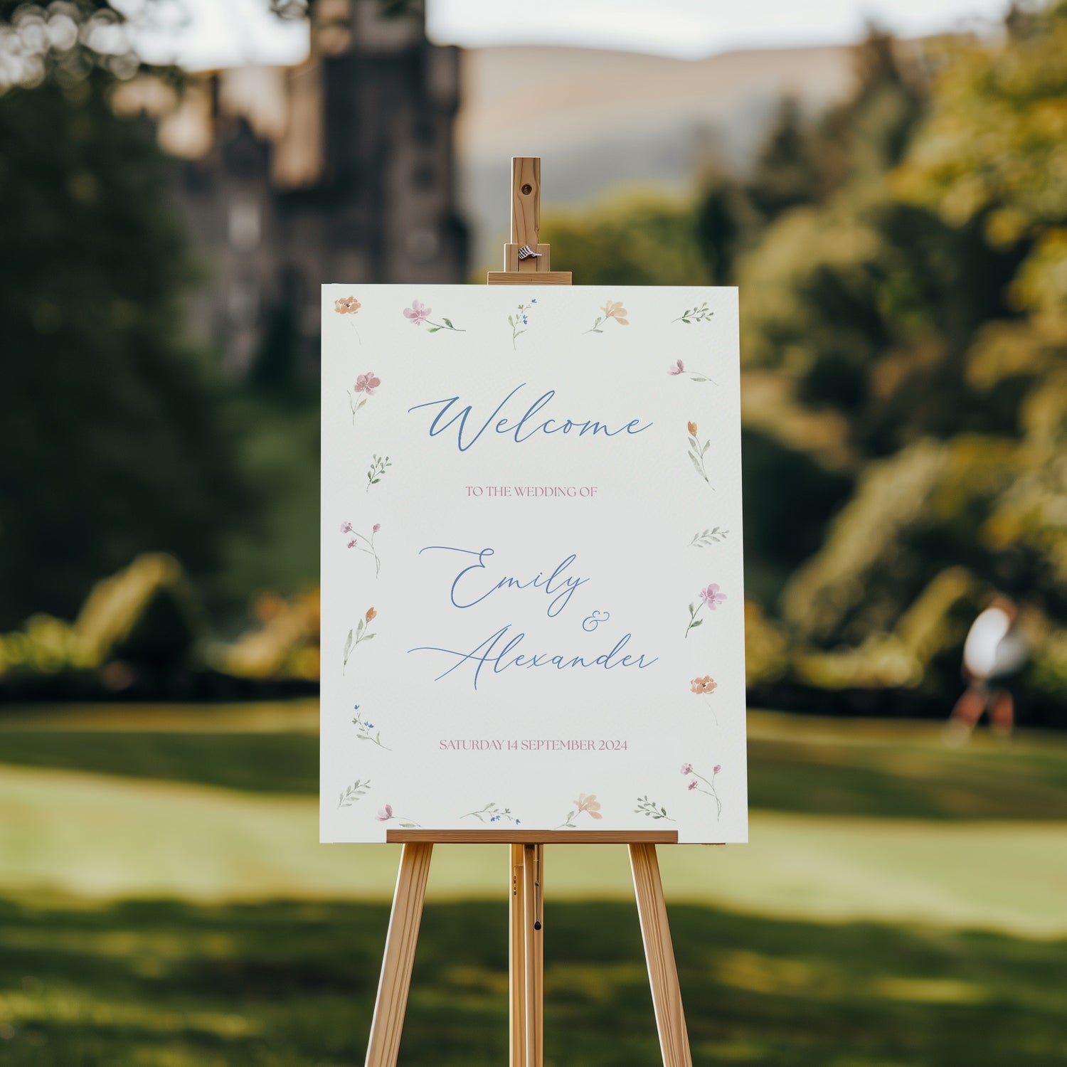 A welcome sign that has a delicate floral border featuring soft pastel-colored flowers and greenery. In the center, "Welcome to the Wedding of Emily & Alexander" is written in an elegant blue script, with the wedding date, "Saturday 14th September 2024," below in a smaller pink font. The overall design exudes a romantic, nature-inspired theme, harmonizing with the natural surroundings.