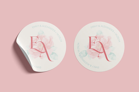 Two versions of a circular wedding sticker featuring the initials "E & A" in a bold pink serif font, set against a soft watercolour floral background. Around the border, the names "Emily & Alexander" and the date "14.09.24" are written in a light teal serif font. One sticker appears to be peeling slightly at the edge, all placed on a soft pink backdrop.