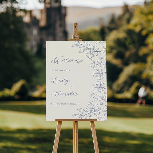 A wedding welcome sign is displayed on an easel in a picturesque garden with a distant castle in the background. The sign features a clean, cream-coloured backdrop with elegant blue floral illustrations along the right-hand side. The text reads "Welcome to the wedding of Emily & Alexander," written in a delicate script font. Below, the wedding date, "Saturday 14 September 2024," is displayed in small, refined lettering.