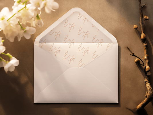 A close-up of a white envelope with a monogram liner featuring the initials "E + A" in a repeating pattern, written in a soft peach script.