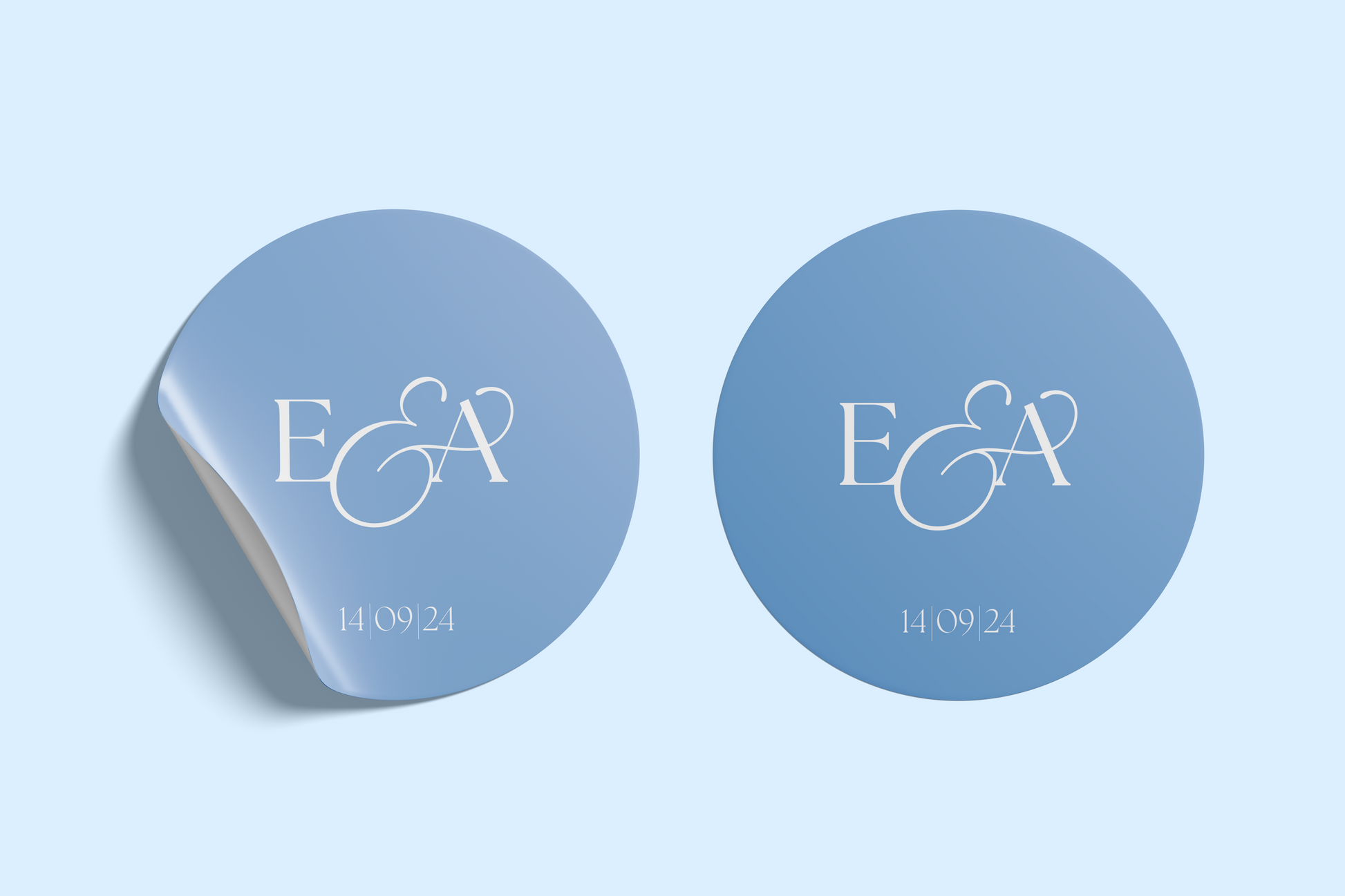 Two versions of a circular wedding sticker featuring the initials "E & A" in an elegant white serif font, with the date "14 | 09 | 24" beneath, set against a soft blue gradient background. One of the stickers appears to be peeling slightly at the edge.