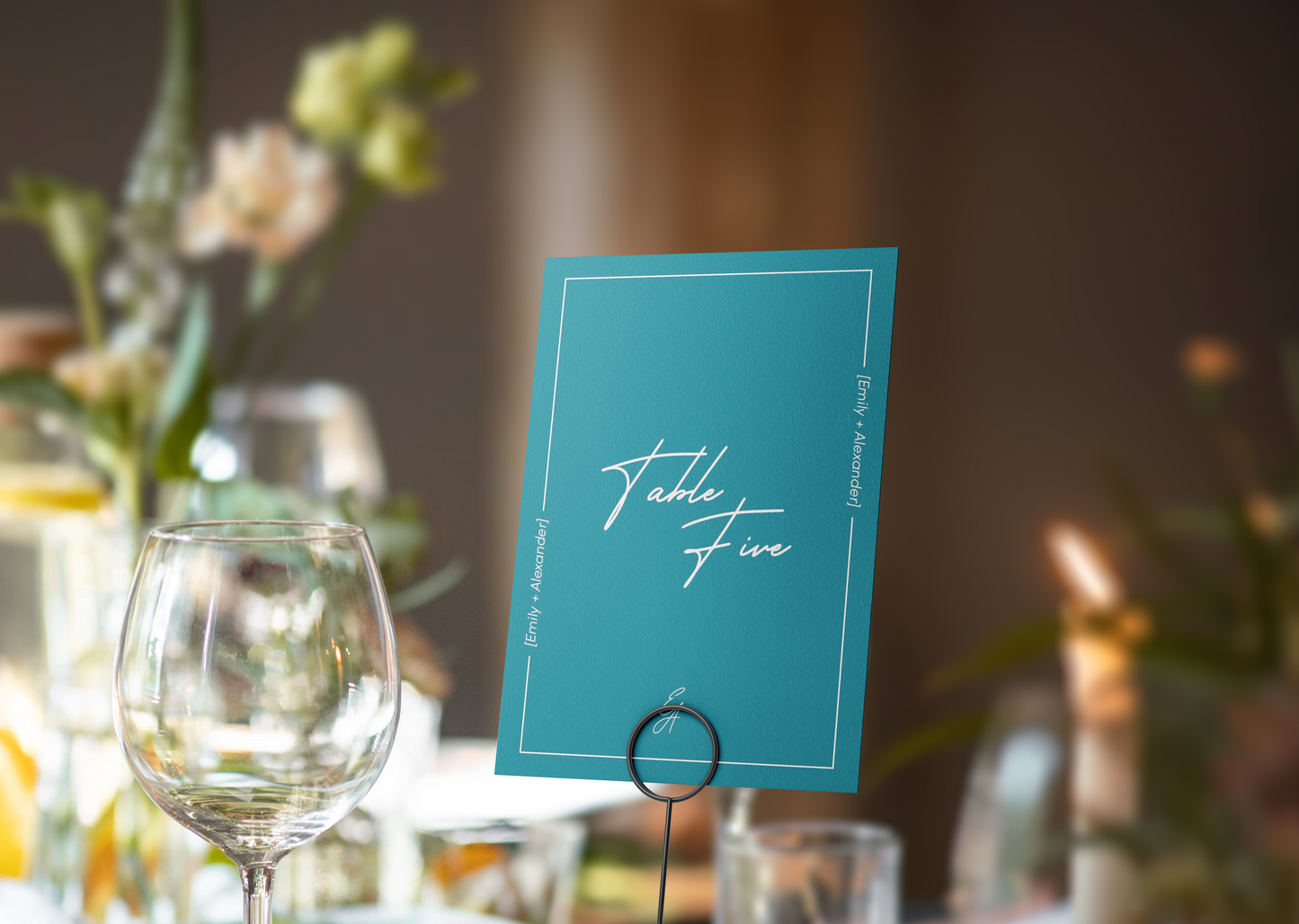 A modern table number card in bold teal, displaying "Table Five" in an elegant handwritten script at the center. The card is bordered by a minimal white frame with the names "Emily + Alexander" written vertically along the sides. 