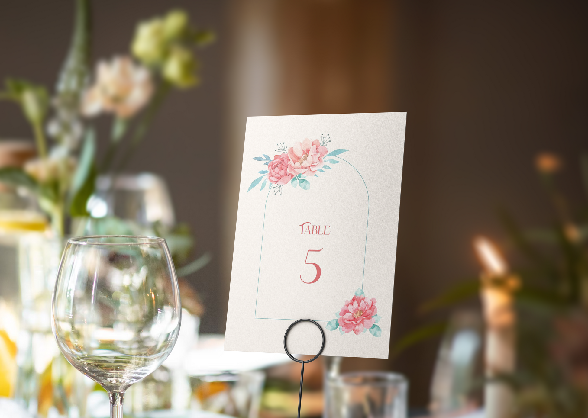 Atable number card set against a sophisticated reception setting. The card displays "Table 5" in an elegant serif font, framed by a delicate arch adorned with soft watercolor flowers in shades of pink and teal at the top and bottom. 